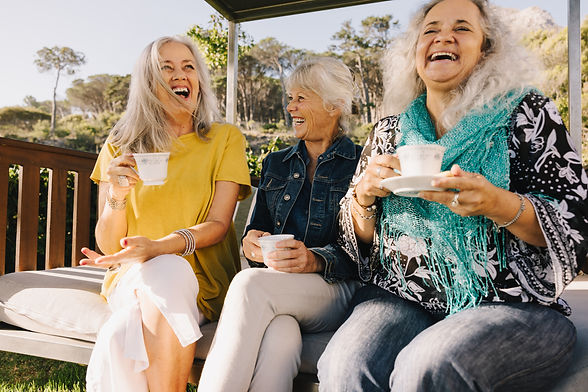 Three 55+ senior women outside enjoying coffee and laughing together seated on a bench