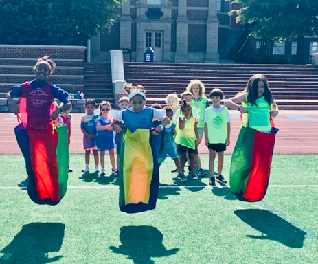 Three children mid-air during a sack race on a green field with others watching behind.