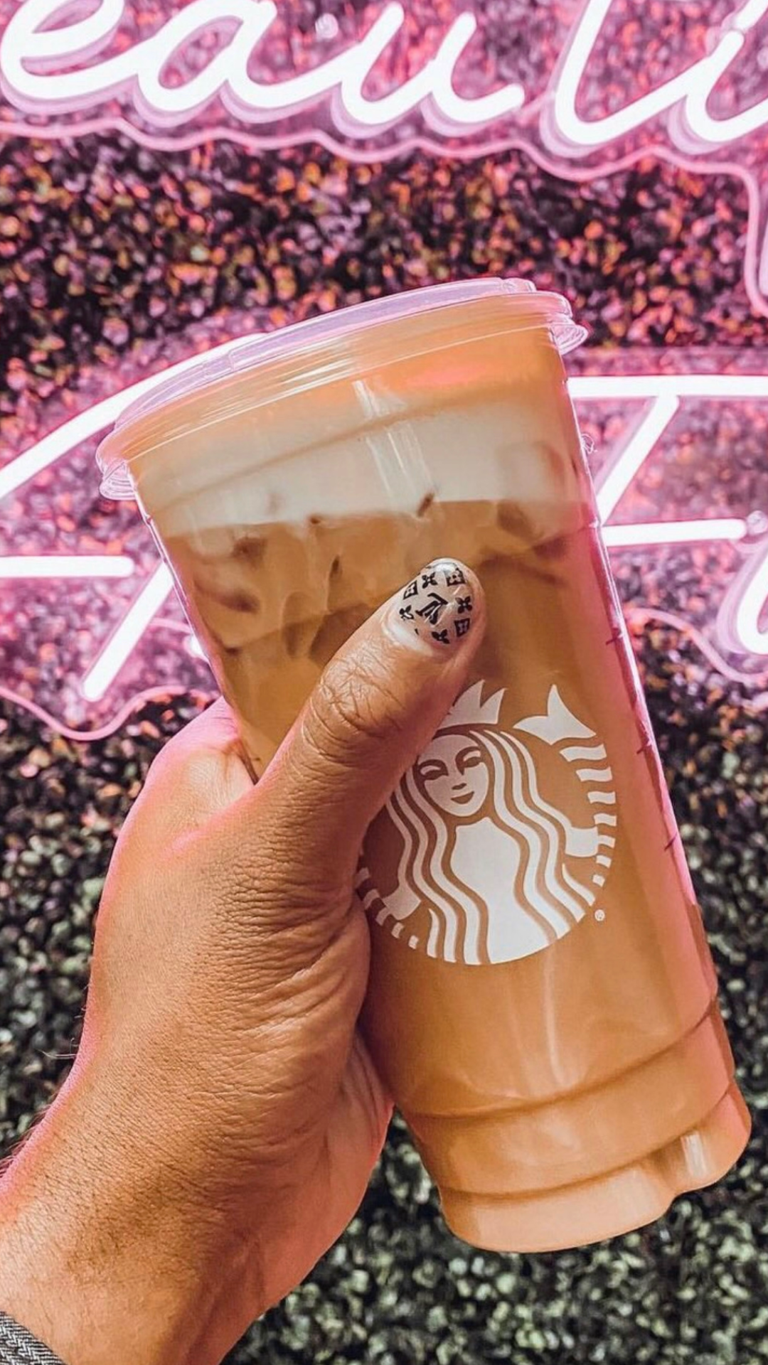 Man holding a Starbucks drink in front of a neon sign with a Louie Vutton inspired patterned nail art on thumb.