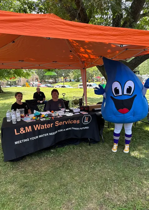 L&M Water Services community outreach booth at a local event, featuring staff members under an orange tent with promotional materials and a friendly water drop mascot.