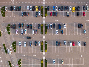 Aerial view of a parking lot with neatly arranged cars, various colors, on a paved surface with marked parking lines and green patches.