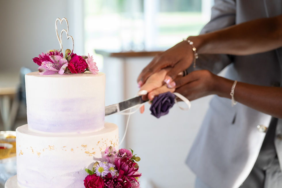 Close-up of a married couple cutting a two-tiered wedding cake decorated with purple and magenta flowers, featuring a heart-shaped cake topper. The couple’s hands are gently holding the knife together, surrounded by soft natural light.