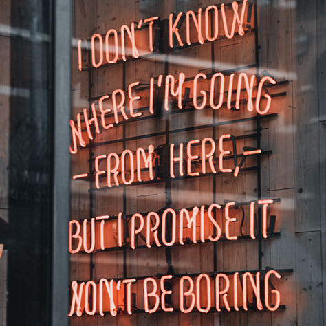 Neon red text on a wooden wall, reads "I don't know where I'm going from here, but I promise it won't be boring." Reflective and moody.