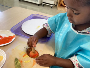 Montessori student in a blue smock painting orange on a leaf with a brush. Paper plates and colorful art supplies on table in a classroom setting.
