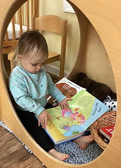 A toddler reads "Little Miss Muffet" in a cozy wooden nook. Dressed in a blue sweater, surrounded by baskets and soft toys, focused and calm.