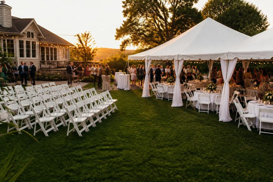 Elegantly set outdoor backyard wedding with white folding chairs arranged in rows, round tables with floral centerpieces under a white canopy tent, and guests mingling in a manicured yard during golden hour