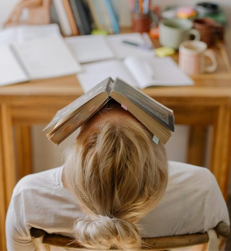 A college student sitting at her dorm desk with her head back and a book opened on her head