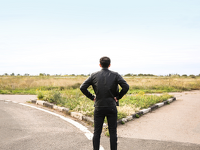 A man wearing dark clothes stands at a crossroads. The rod to the left is paved, the road to the right is made of dirt.