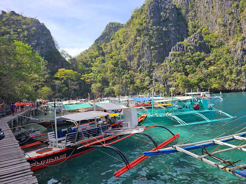 Boats docked near a wooden walkway with tourists in orange life vests. Limestone cliffs and green trees surround the clear blue water. In Kayangan Lake Coron