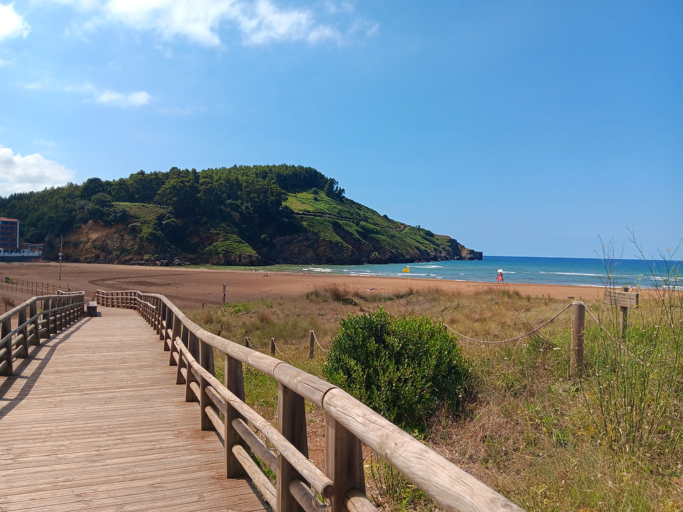 Wooden boardwalk leads to a scenic beach with a green hill in the background. Blue sky and calm ocean create a serene atmosphere.