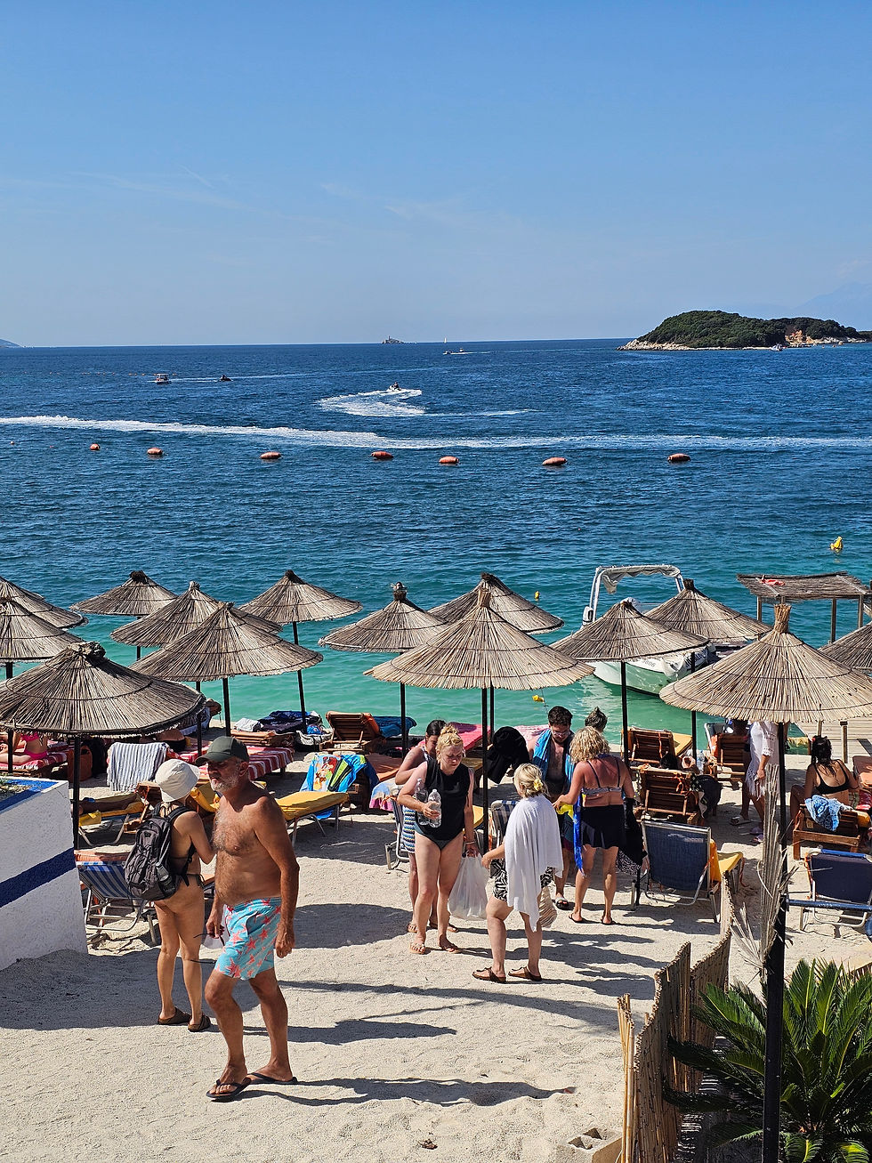 Beach scene with people in swimwear under straw umbrellas. Blue sea and sky in background. Boats and distant island visible. Relaxed mood.