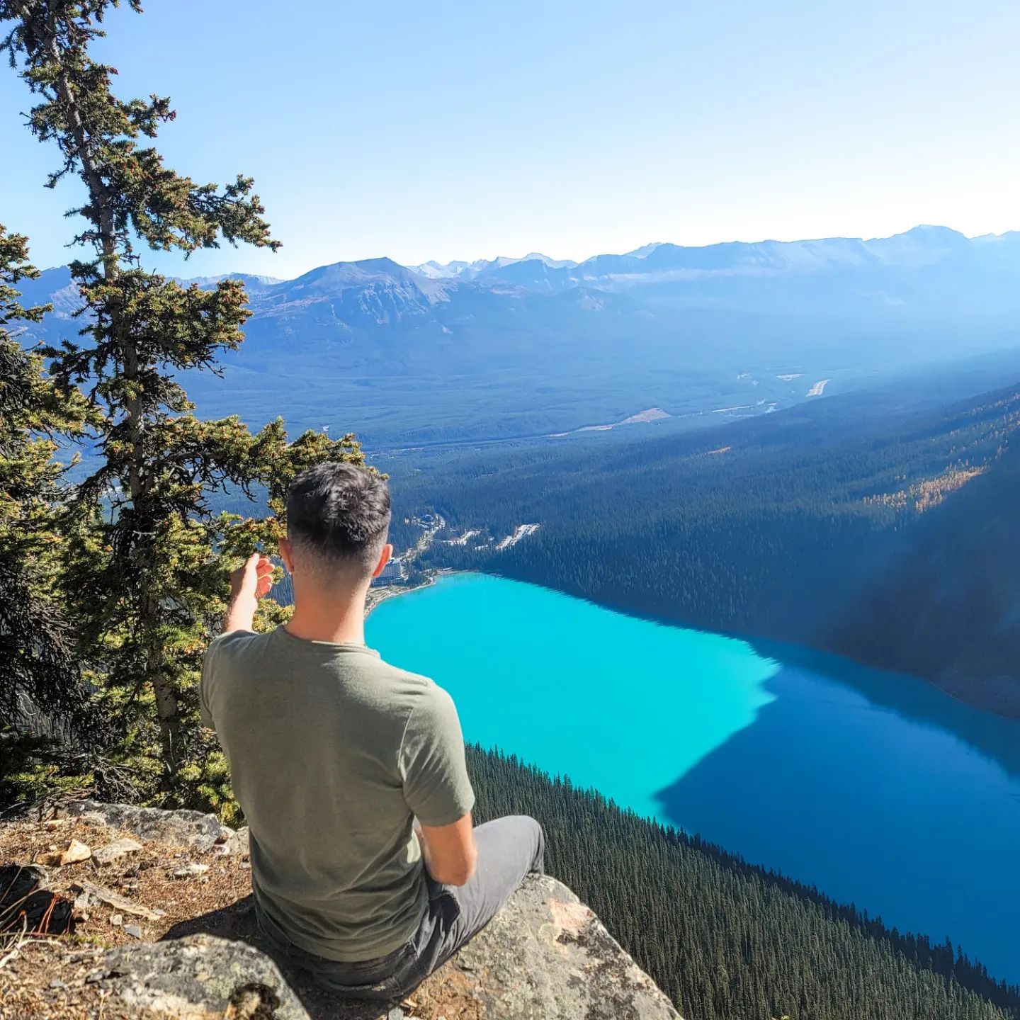 Person sitting on a cliff, pointing towards a turquoise lake below, surrounded by mountains and trees under a clear blue sky.