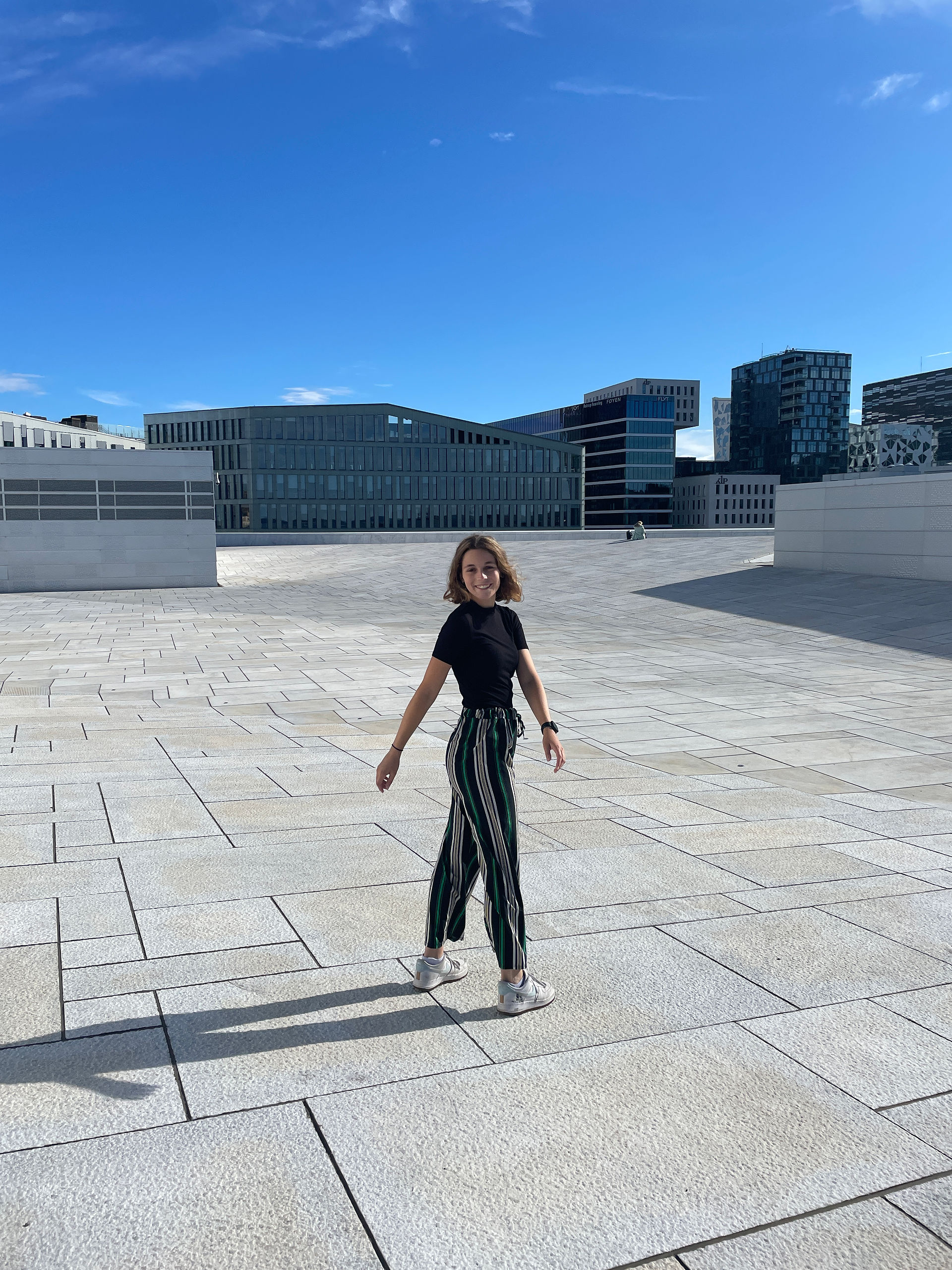 A girl with green striped pants and a black tshirts smiling to the camera with the opera house on the background