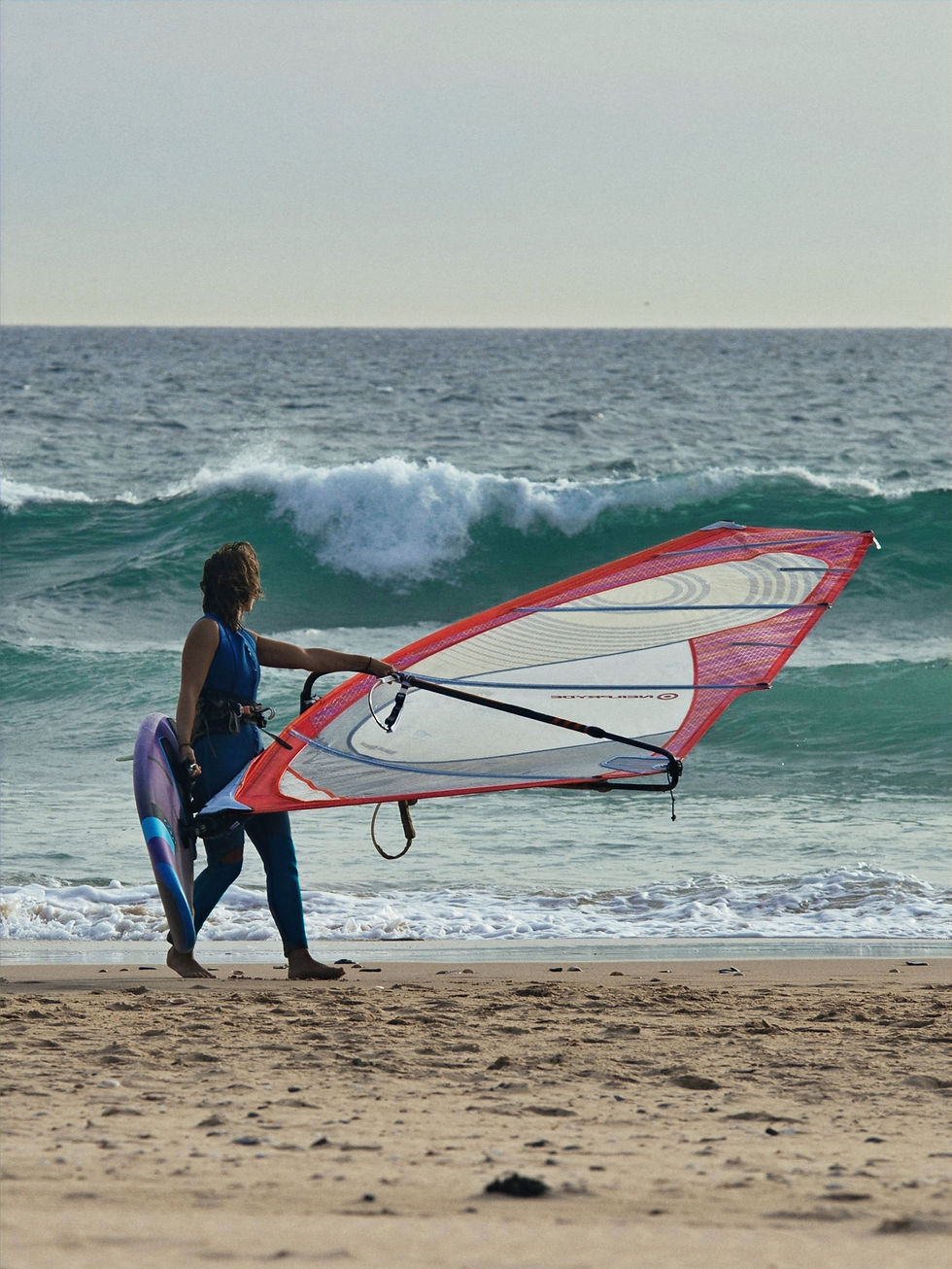 Person carrying a windsurfing board and full sail rig on the beach