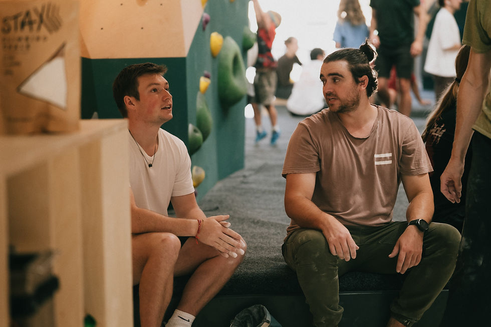 two guys sit together on the bouldering mats talking, they seem to be in conversation. the guy on the left wears a white tshirt with hands clasped together with chalk. Theguy on the right wears a mocha coloured t-shirt and olive green pants, and has a top knot.