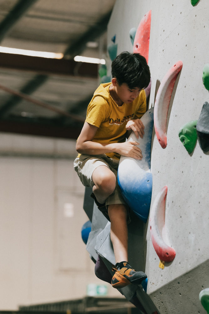 Young asian male climber with both hands gripping a large blue bouldering hold. His body is crunched tightly around the hold and he is looking into the hold where his hands are.