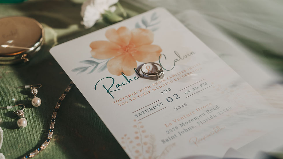 Wedding invitation with floral design, rings, pearl earrings, and diamond bracelet on green fabric. Sunlight creates a romantic mood.