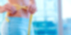 Woman measuring waist with a tape measure. She is wearing blue shorts. The background is a blue glass window.