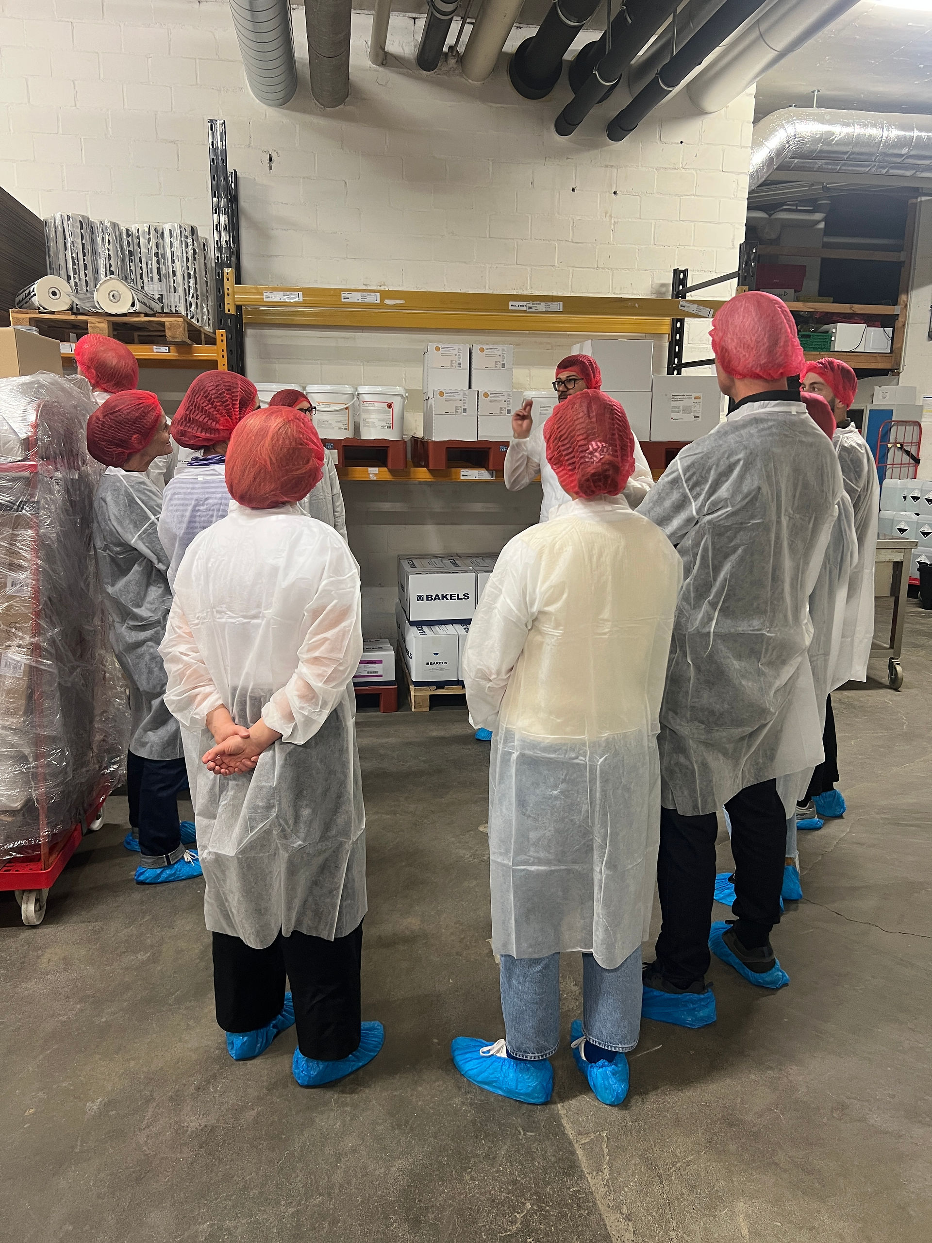 Gruppe von Personen in weissen Schutzkitteln und roten Haarnetzen hört in einem Lagerraum einem Mitarbeitenden zu, der etwas erklärt.