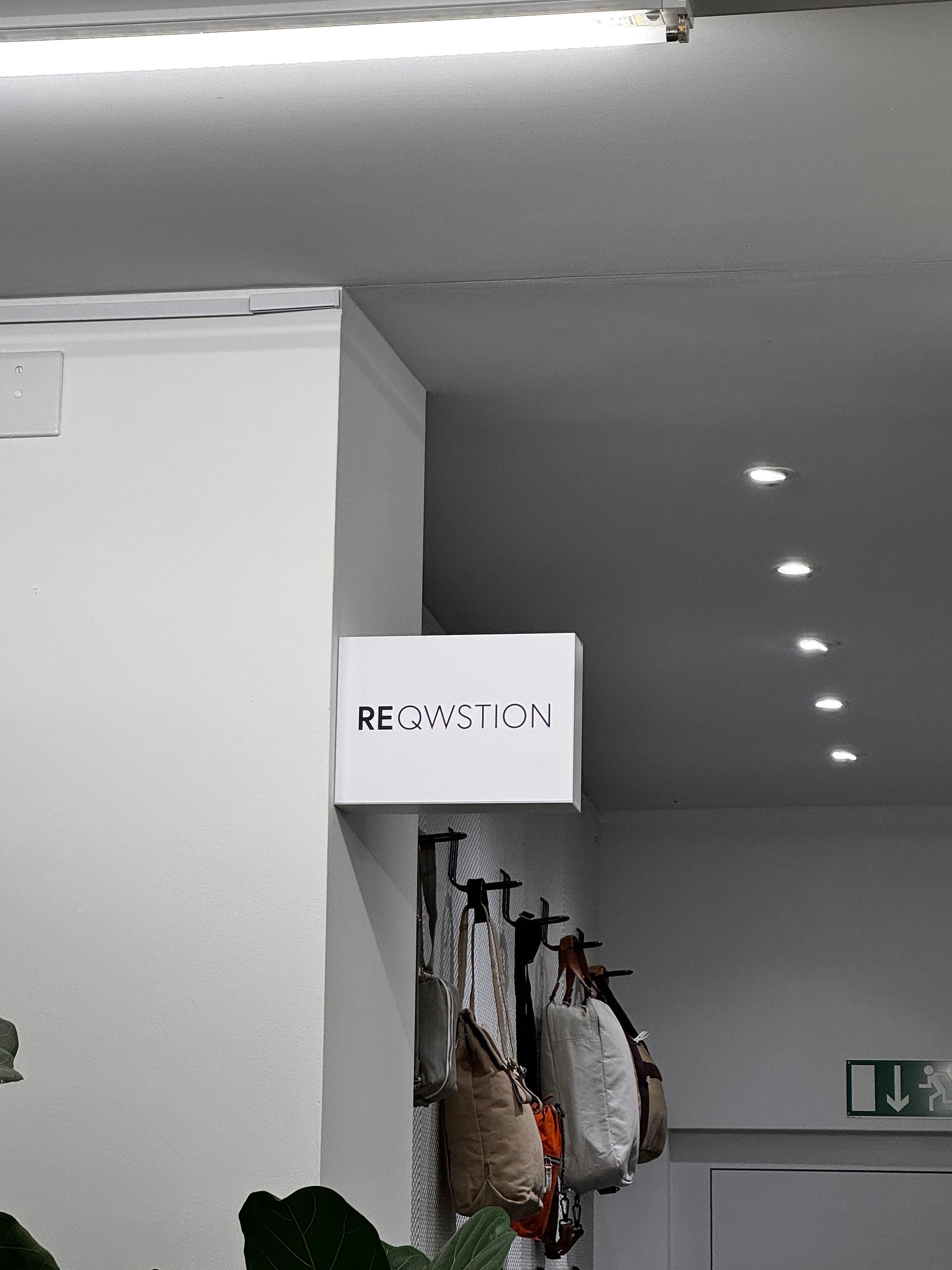 Weisses Leuchtschild mit dem Schriftzug „REQWSTION“ an einer weissen Wand, daneben hängen mehrere Taschen an Haken.
