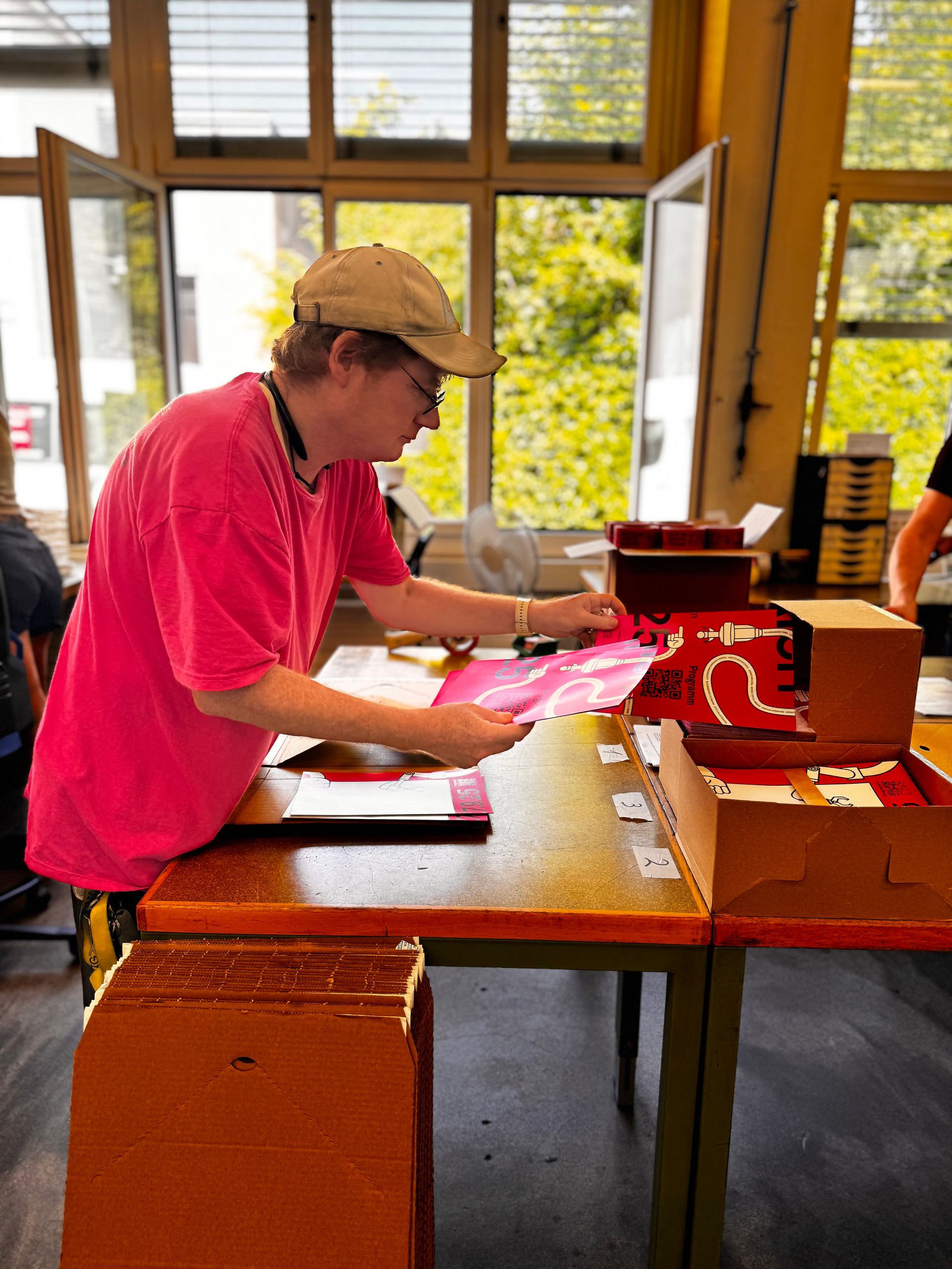 Ein Mitarbeiter der Stiftung Züriwerk in pinkem T-Shirt und beige Cap verpackt pinke Plakate für den Tag der urbanen Produktion an einem Arbeitstisch.