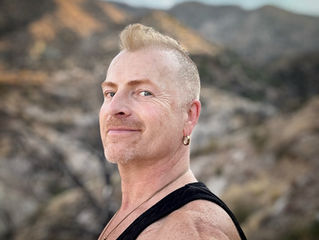 Smiling person with short hair in a black tank top stands against a mountainous, blurred background. Sky is clear with a few clouds.