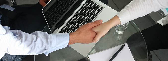 two hands shaking over a desk with a laptop and glass of water