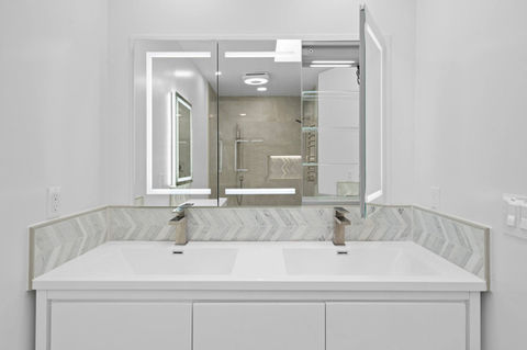 Contemporary bathroom remodel with a double-sink vanity, chevron tile backsplash, and an LED mirror cabinet partially open to reveal sleek glass shelving.