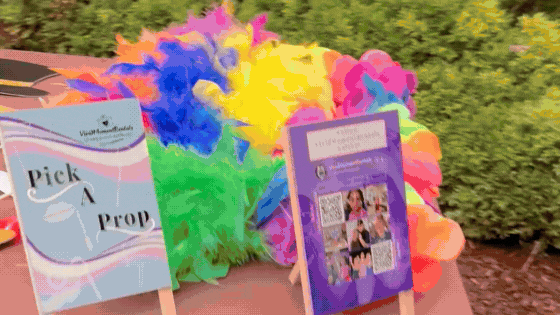 Colorful feather boas on a table with greenery. Two signs read "Pick a Prop" and display images and a QR code. Vibrant, festive atmosphere.