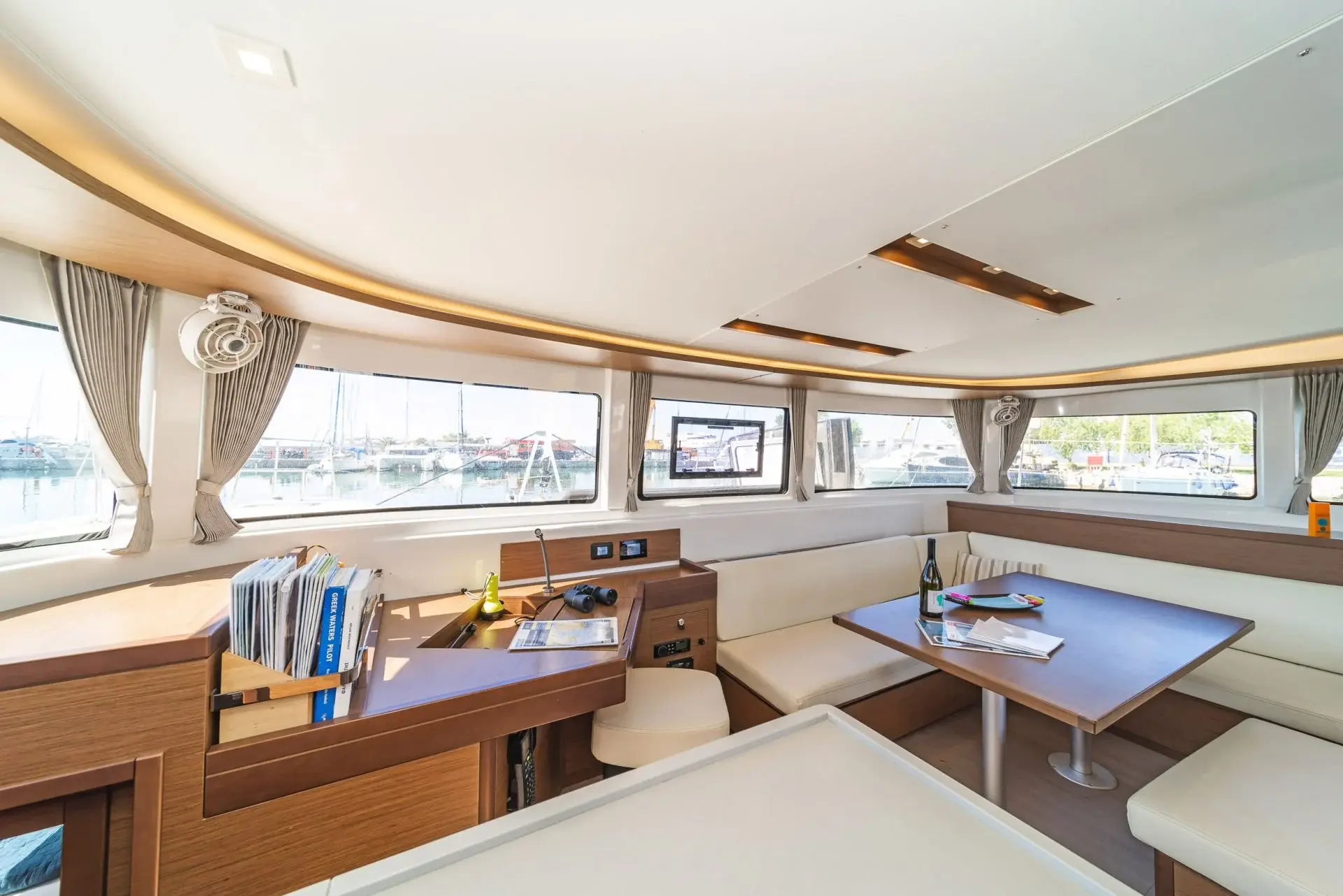 Bright catamaran saloon with panoramic windows, U‑shaped seating, large table, open and airy interior.
