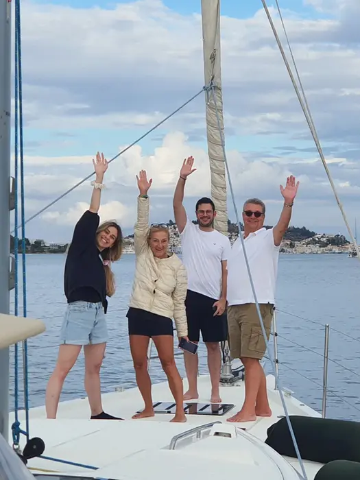 Joyful family waving on the bow, approaching Poros, Family Holiday sailing adventure.