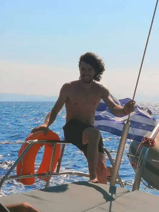 Epic Voyage guest relaxing on starboard pulpit, enjoying day sail between Cyclades islands with turquoise Aegean Sea behind.