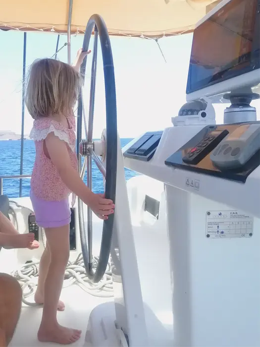 Young girl confidently steering a catamaran, wheel larger than her, unforgettable Family Holiday sailing experience.
