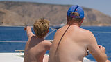 Father and son sitting on forward deck of a catamaran, boy pointing left toward shore, blue Aegean Sea background, Family Holiday sailing in Greece.