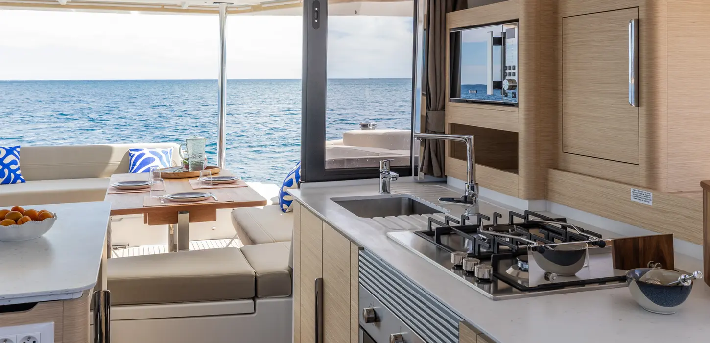 Galley of Fontaine Pajot 44 with stove in foreground, island counter, and bright outside seating area visible beyond, onboard the Family Holiday sailing experience.