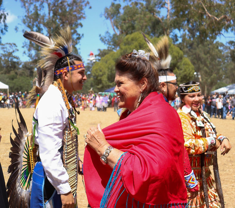 Decorative: Karen and two powwow dancers standing. Karen wears traditional-wear. A man wears fancy dance regalia and the woman wears powwow regalia.