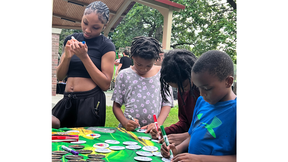 Children sit at a table coloring circular paper designs used to create custom buttons, with adults nearby at an outdoor community event.