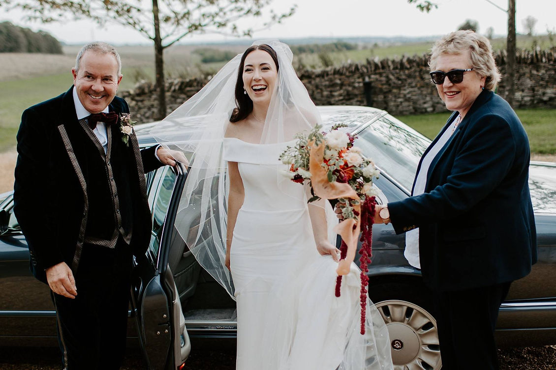 A joyful bride in a white gown and veil stands by a car holding a vibrant bouquet,