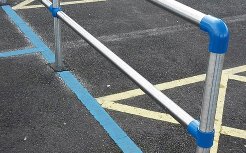 The image shows an outdoor tubular safety railing or barrier system installed next to an asphalt surface