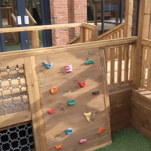 Outdoor wooden play structure featuring an inclined climbing wall with colorful handholds next to a cargo net