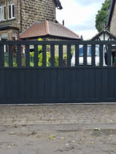 A large, modern black gate blocking a driveway leads to a stone house with a red roof