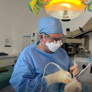A male dentist in blue scrubs and a surgical mask examining a patient under a bright overhead light