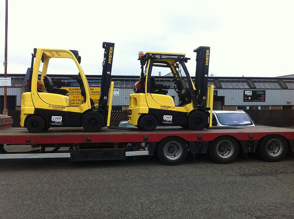 Two yellow Hyster forklift trucks being transported on a red flatbed lorry trailer on a street