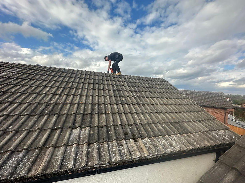 Roofer working on a large, old, grey tiled roof covered in moss and lichen under a cloudy sky