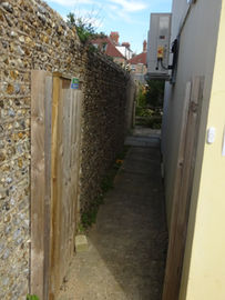 Narrow concrete walkway between a high flint wall and a building with a timber gate