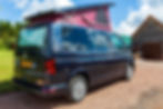 A dark blue Volkswagen campervan parked on gravel with its burgundy pop top roof