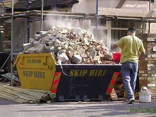 Yellow and black skip overloaded with rubble and bricks, a person standing nearby
