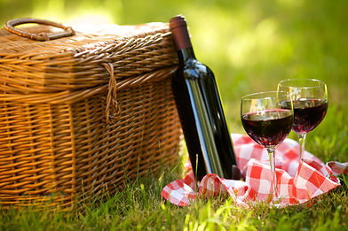 Wicker picnic hamper next to a bottle of red wine and two glasses on red gingham in grass