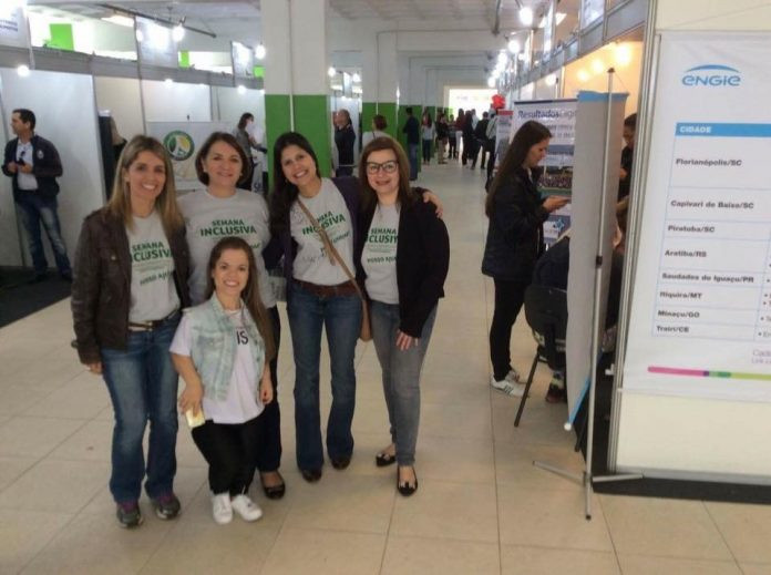 #ParaTodosVerem A imagem é uma foto tirada na Semana Inclusiva de Florianópolis de 2017. São retratadas ao centro 05 mulheres brancas sorridentes, sendo que quatro de estatura mediana usando a camisa do evento se posicionam atrás e uma com nanismo na frente. Ao redor, vemos vários estandes onde ocorrem as atividades do evento, e várias pessoas aparecem. Na camisa do evento lê-se “Semana Inclusiva” em letras verdes. A entrevistada Luciana Sans é a segunda à direita.