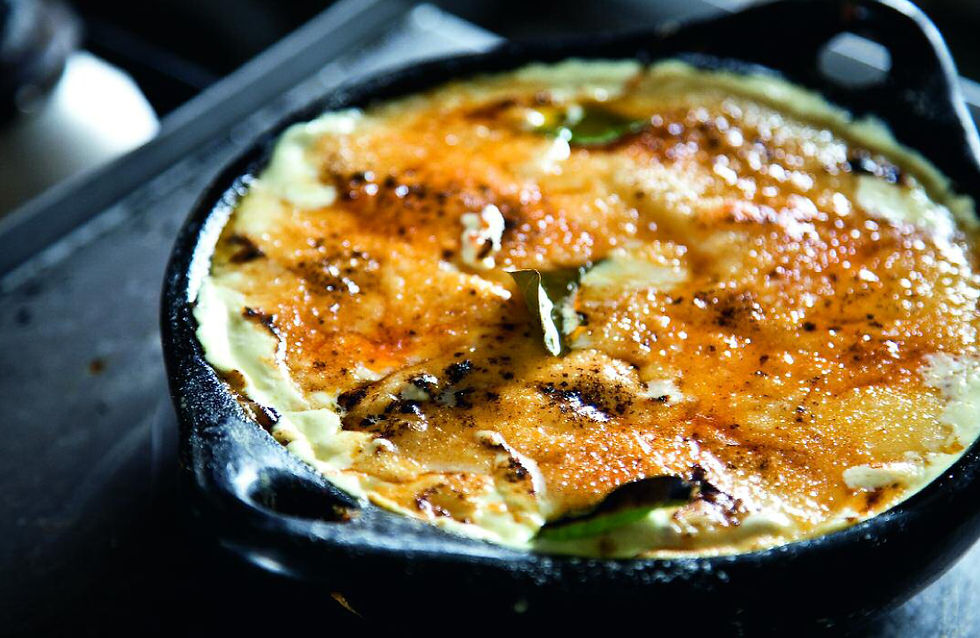 Golden baked dish in a black skillet with speckled top, garnished with herbs. Set on a dark countertop with a blurred background.